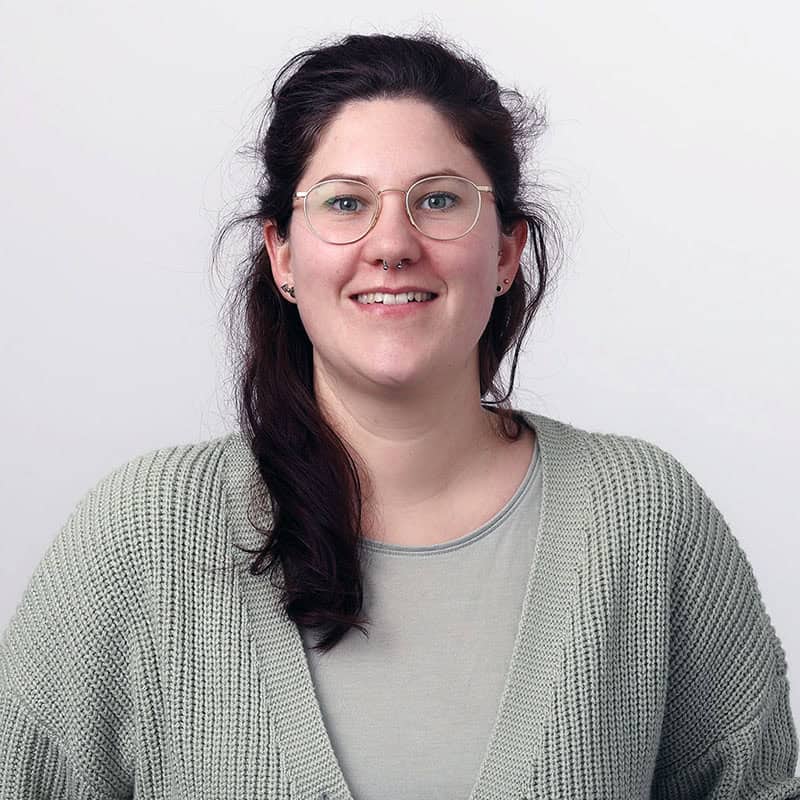 Junge Transfrau mit Brille und dunklem Haar, lächelnd, im weißen Hintergrund.