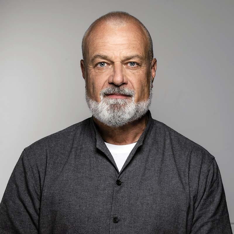 Freundlicher älterer Mann mit grauem Bart und blauen Augen, trägt dunkles Hemd, neutraler Hintergrund, modern und professionell.