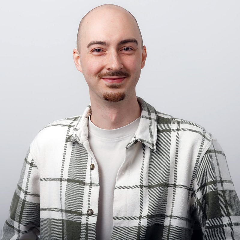 Junges Mannbild, lächelnd mit Glatze und Bart, in weißer Hemdjacke vor hellem Hintergrund.