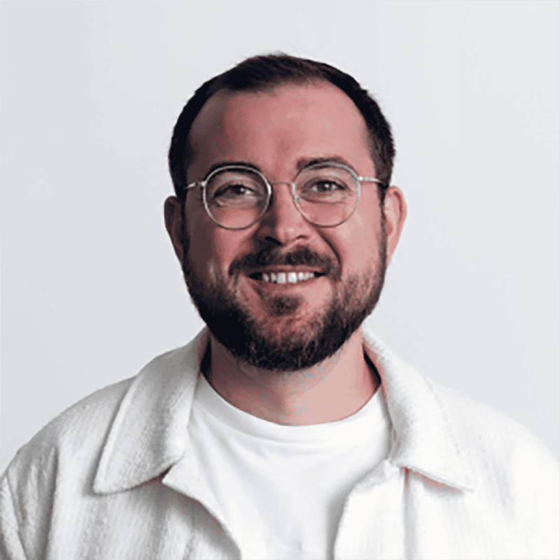 Freundlicher schwuler Mann mit Brille und Bart, lächelnd, im weißen Pullover vor neutralem Hintergrund.